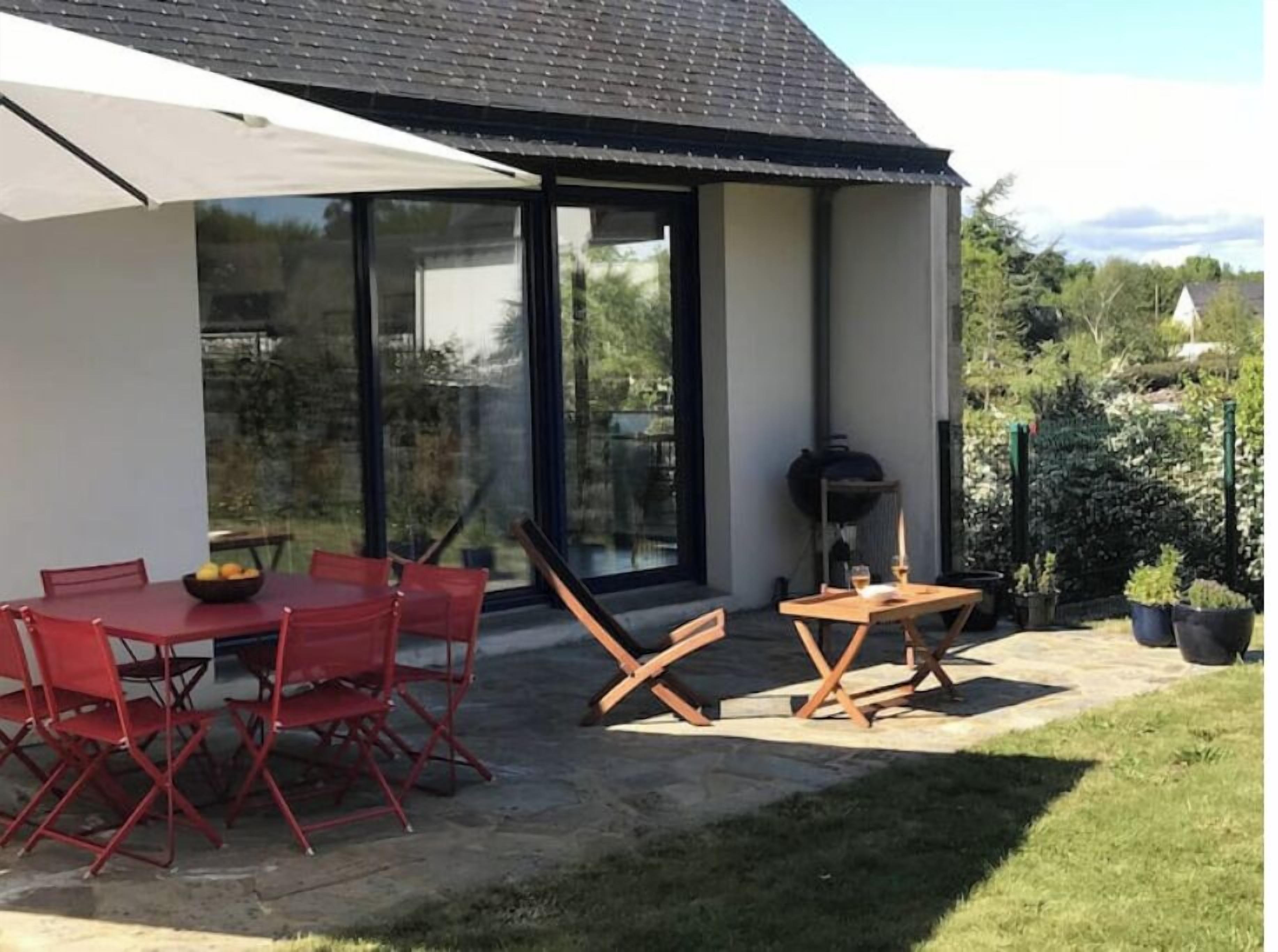 A modern house with a patio area featuring a dining table and chairs, a barbecue grill, and a large umbrella.