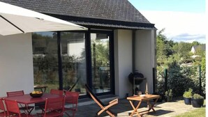 Ett modernt hus med en uteplats som har ett matbord och stolar, en grill och ett stort parasoll.