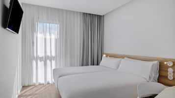 A modern hotel room with a large bed, a TV mounted on the wall, and a window with sheer curtains.