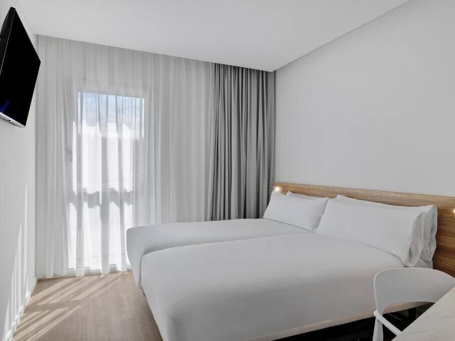A modern hotel room with a large bed, a TV mounted on the wall, and a window with sheer curtains.