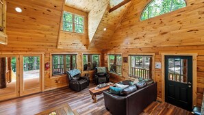 Interior de una cabaña de madera con piso de madera, un sofá de cuero, una mesa de centro y ventanas amplias que ofrecen vistas del bosque circundante.
