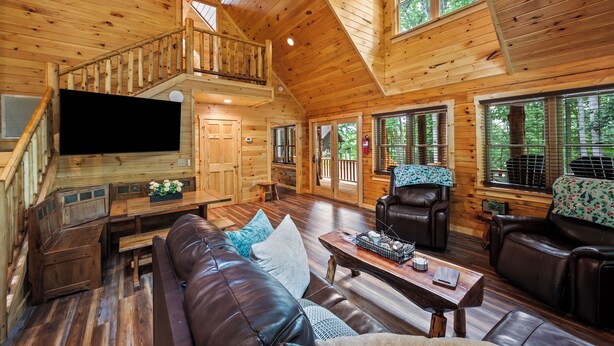 Una cabaña de madera espaciosa con habitación en el altillo, sala de estar con mobiliario de cuero y amplias ventanas que ofrecen vistas al bosque circundante.
