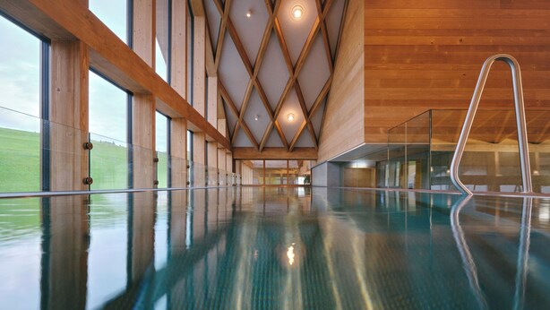 Une grande piscine intérieure avec des poutres en bois et des murs en verre, une échelle en métal et une vue sur un champ verdoyant à l’extérieur.