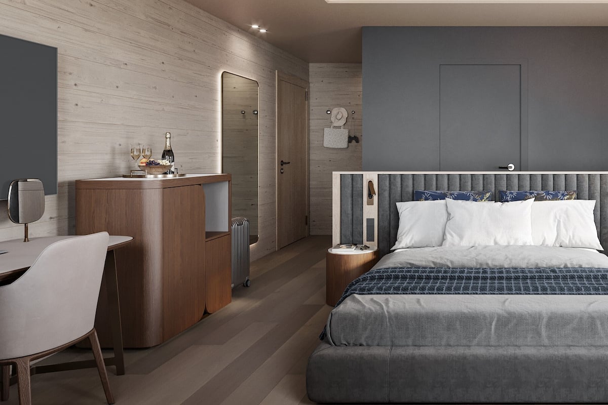 A modern hotel room with a wooden desk, a bed with a grey and blue patterned bedspread, and a grey headboard.