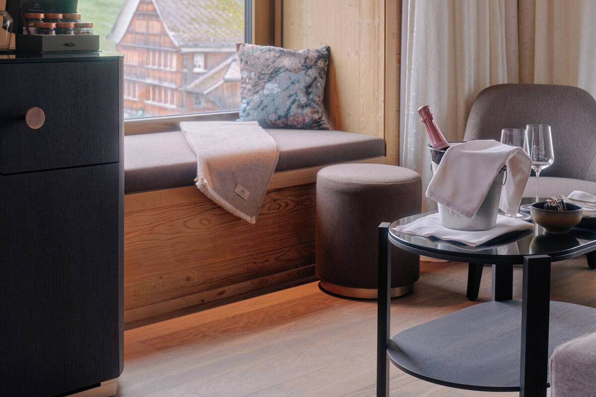 A room with a wooden interior, a window with a view of a house and green landscape, a sofa with a cushion, a small table with a glass and a bottle, and a cabinet.