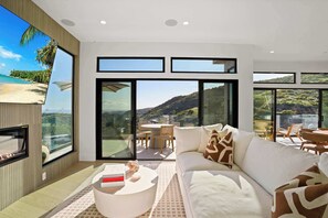 A modern living room with a large sofa, a coffee table, and a TV. There are large windows offering views of a beach and hills. - Samara by Stay Awhile Villas (Malibu)
