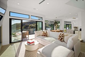 A modern living room with a large sectional sofa, a round coffee table, and a dining area with a table and chairs. Large windows provide a view of the outdoors. - Samara by Stay Awhile Villas (Malibu)