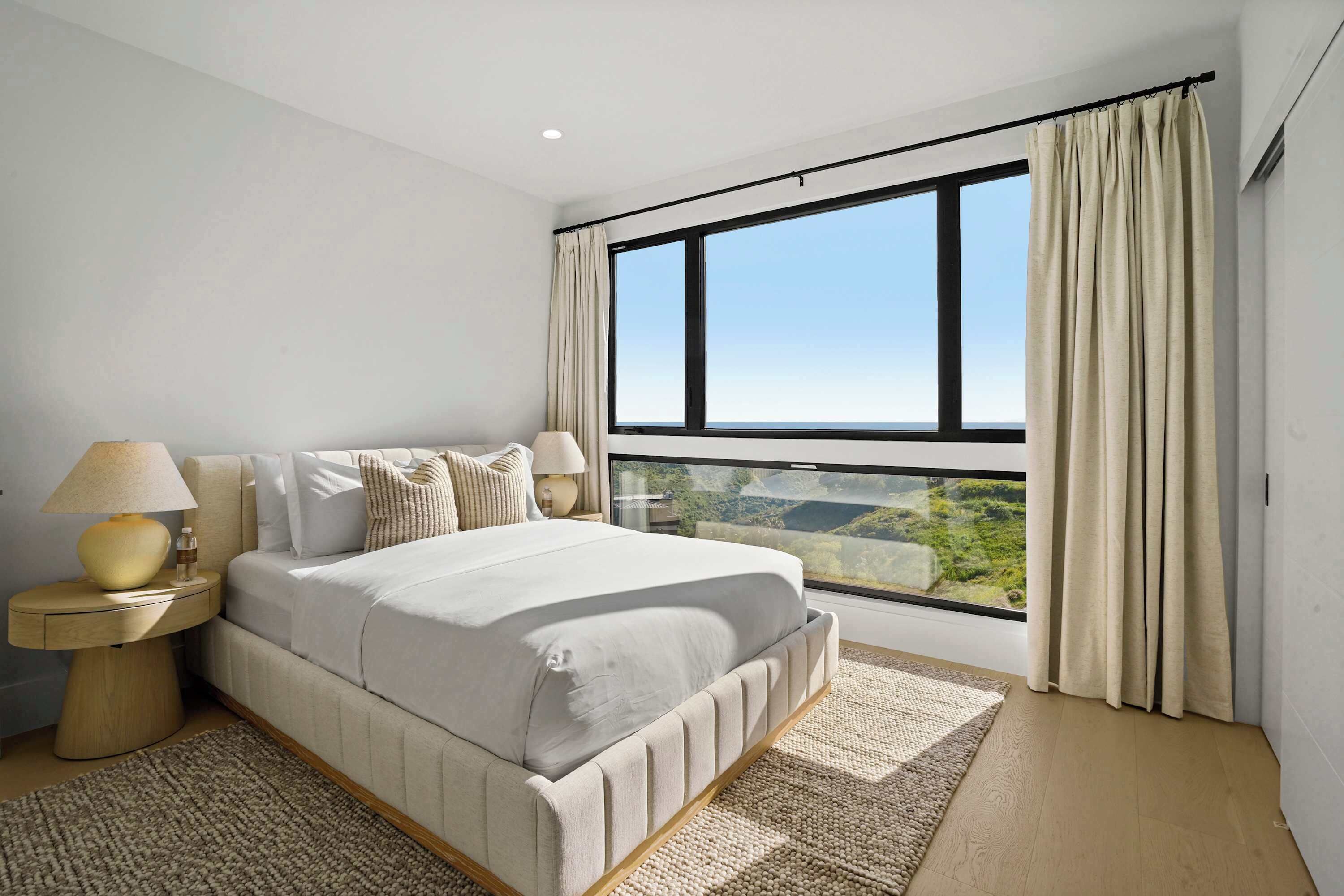 A bedroom with a large window, a bed with a headboard, two bedside tables with lamps, and a view of the outdoors.