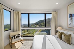 A hotel room with a large bed, a chair, and a view of a mountainous landscape through large windows. - Samara by Stay Awhile Villas (Malibu)