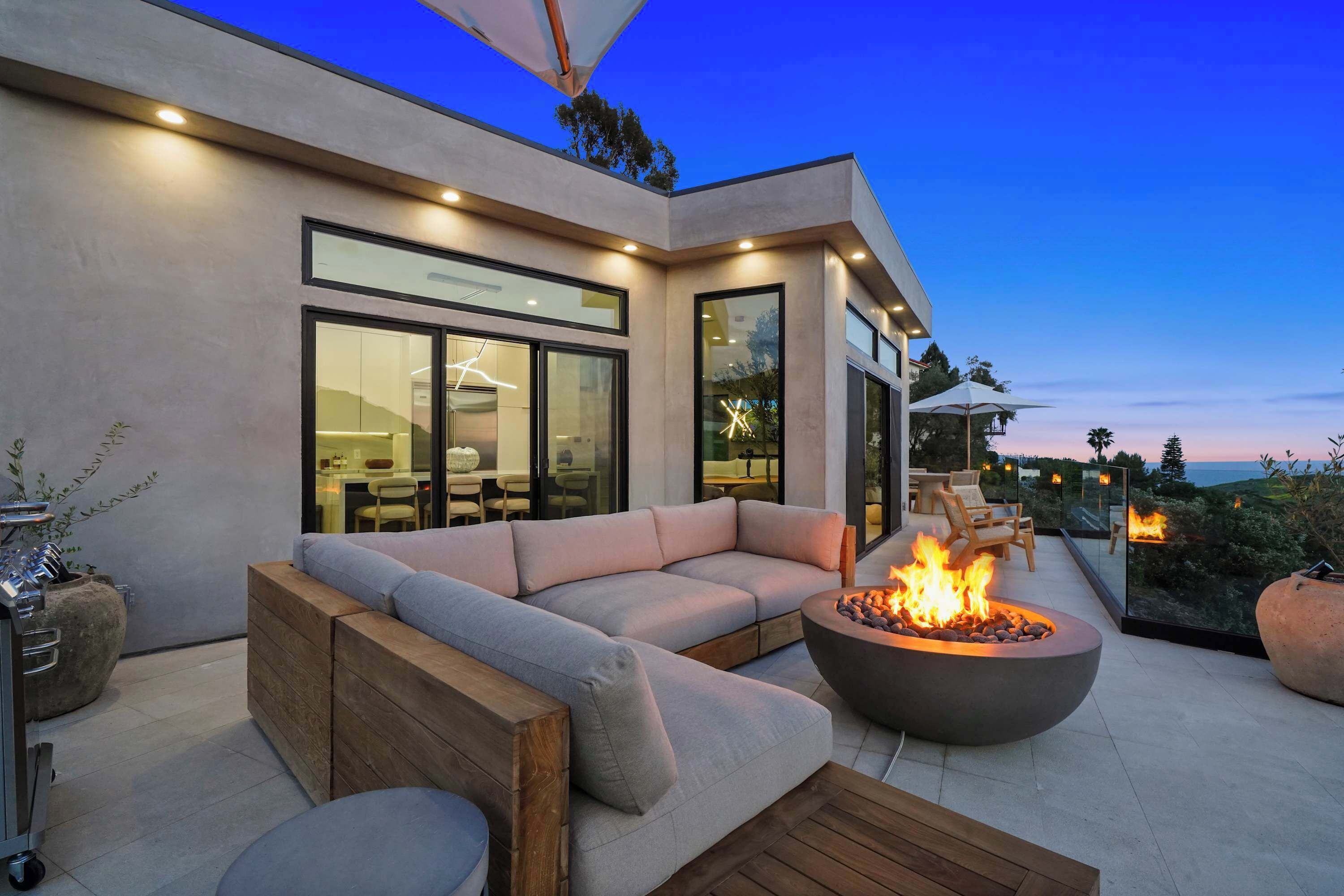 A modern outdoor living area with a sectional sofa, a fire pit, and a dining area visible through large windows.