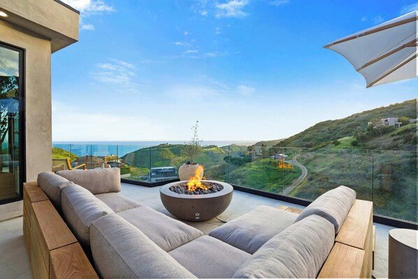 A modern outdoor seating area with a fire pit, overlooking a scenic hillside and ocean view.