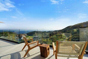 A balcony with wooden chairs and a small table, offering a view of a mountainous landscape through a glass railing.