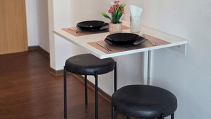 Un petit coin repas avec une table blanche, deux tabourets noirs, une assiette noire, un petit vase avec des fleurs roses et un récipient en verre transparent.