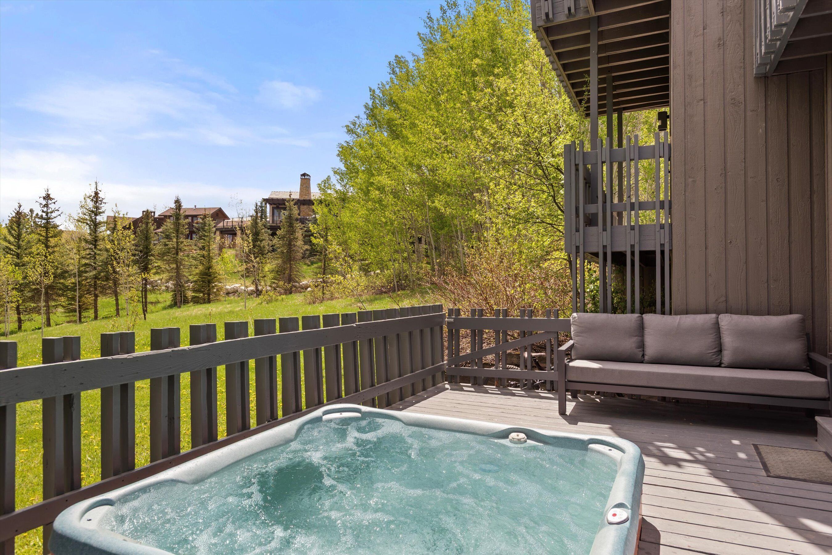 A wooden deck with a hot tub, a gray sofa, and a view of a forested area.
