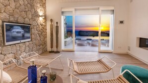 Un moderno salón con pared de piedra, televisor, sillones y vista a la puesta de sol sobre el mar.