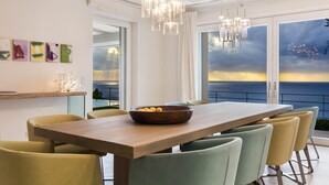 Un comedor con una mesa de madera, sillas y un cuenco de fruta. Hay ventanas amplias con vistas al océano y al atardecer.
