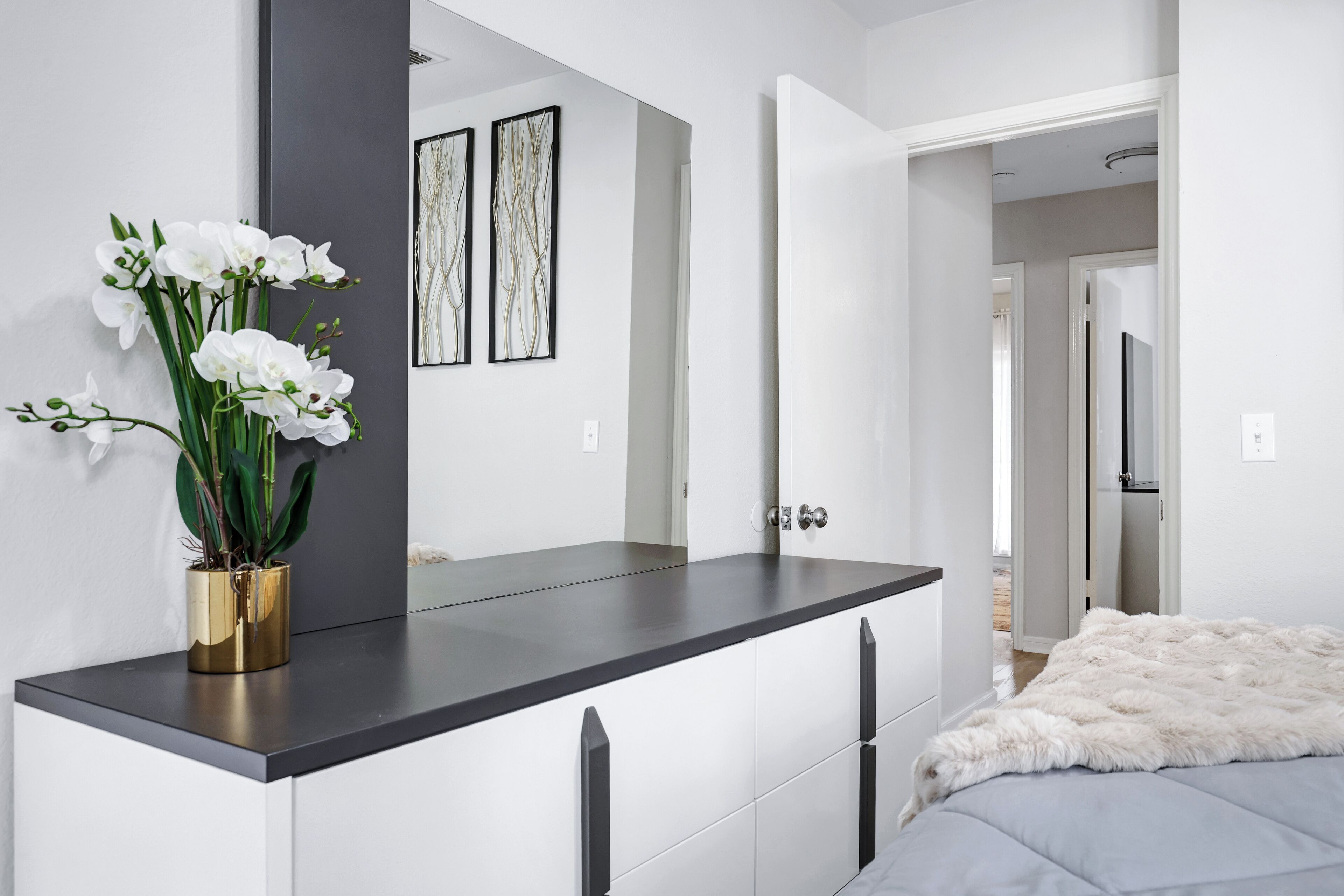 Een moderne slaapkamer met een grijs bed, een witte, zachte deken, een gouden vaas met witte bloemen en een grote spiegel op een donkere dressoir.