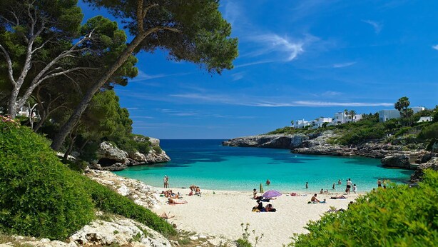 A beach with clear turquoise water, people sunbathing, and white buildings on a hillside.