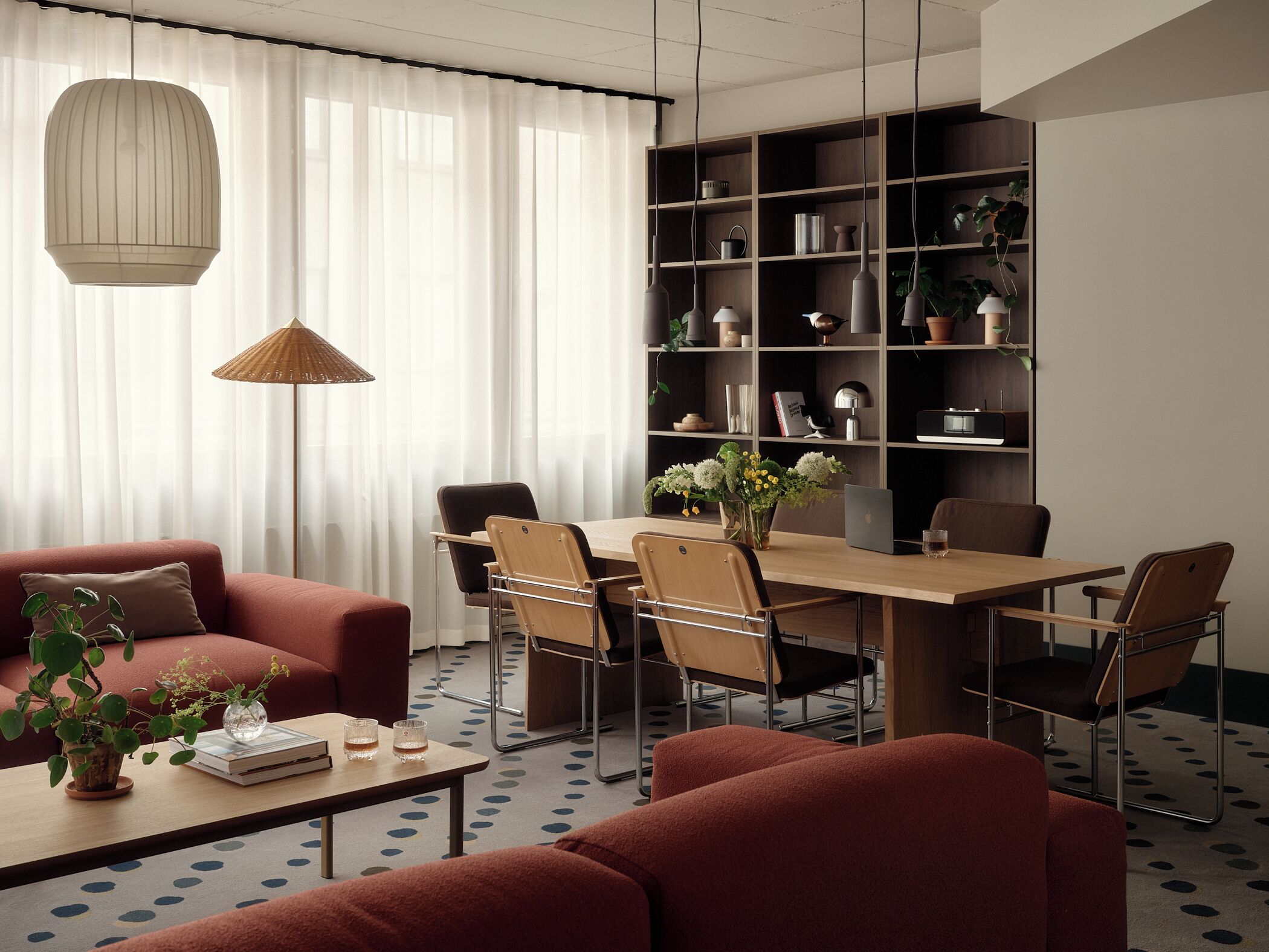 Ein modernes Wohnzimmer mit einer Couch, einem Couchtisch und einem Esstisch mit Stühlen. Ein Bücherregal mit verschiedenen Gegenständen und eine Hängelampe sind ebenfalls vorhanden.