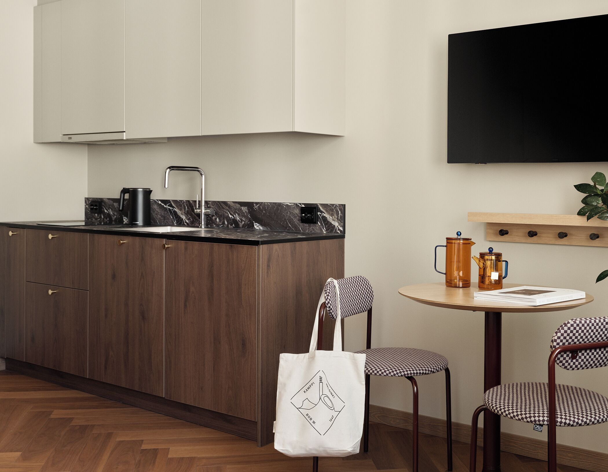 A modern kitchen with dark wooden cabinets, a black marble countertop, and a white kettle. There is a round wooden table with two checkered chairs and a coffee pot. A white tote bag is placed on one of the chairs.