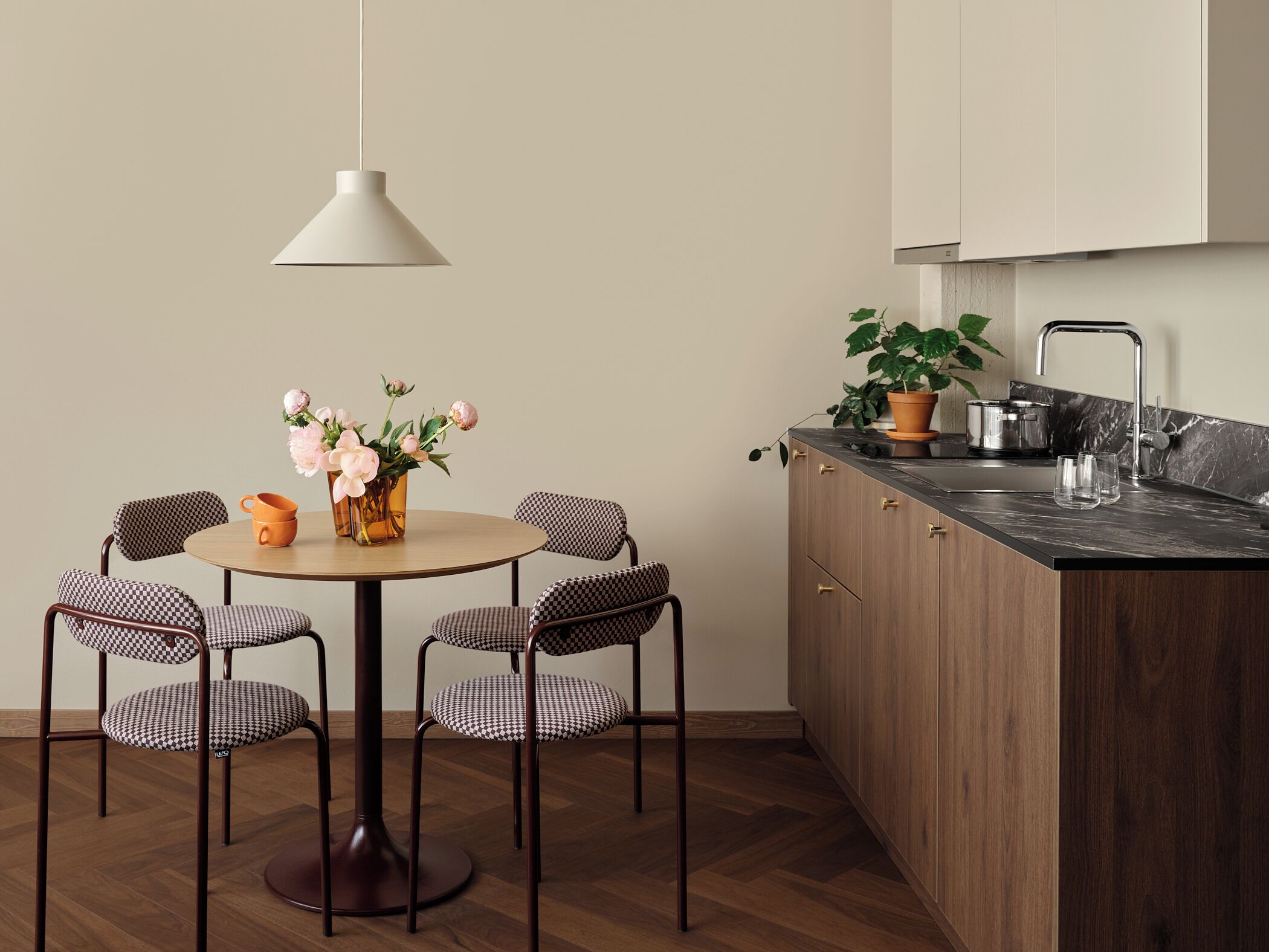 A modern kitchen with a round dining table, checkered chairs, a hanging light, and a potted plant.