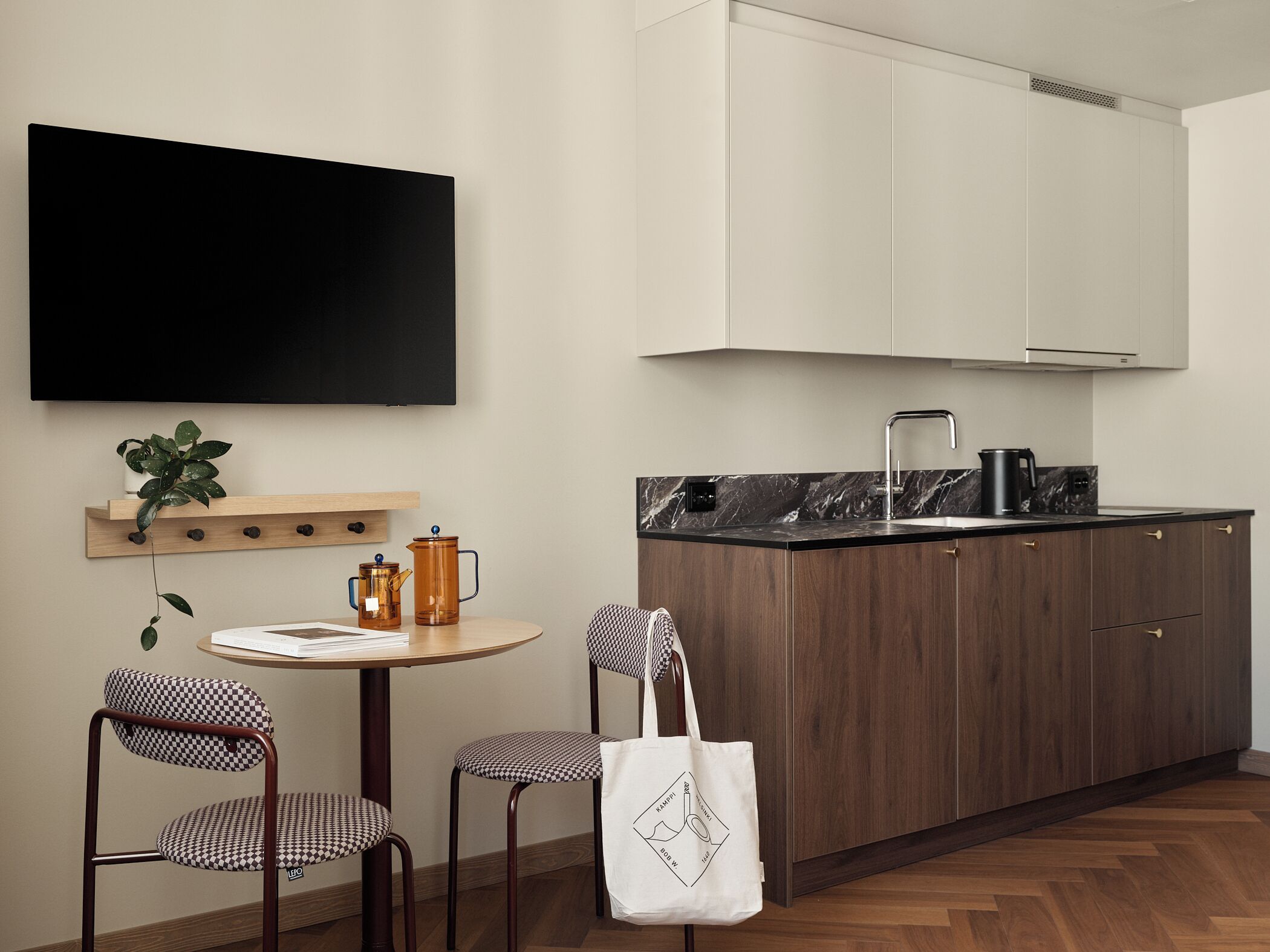 A modern kitchen with wooden cabinets, a black countertop, and a built-in stove. There are two chairs with checkered cushions and a small table with a plant and a kettle.