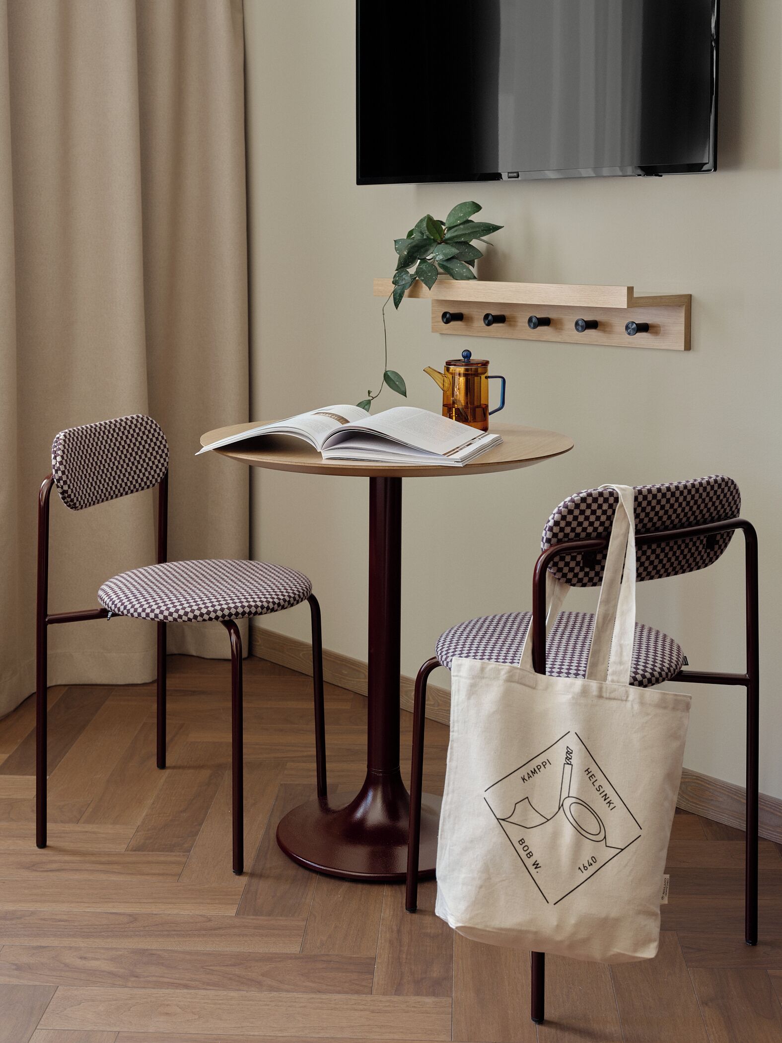 A small, cozy room with a round wooden table, two checkered chairs, a single hanging plant, a mounted television, and a wooden shelf with hooks.