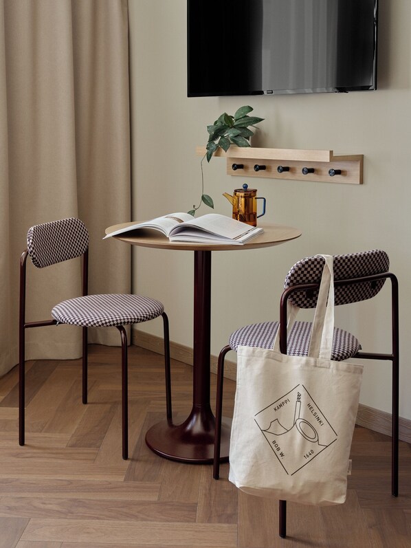 A small, cozy room with a round wooden table, two checkered chairs, a single hanging plant, a mounted television, and a wooden shelf with hooks.