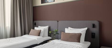 A hotel room with two single beds, a large window with curtains, a wall-mounted TV, and a framed picture above the beds.