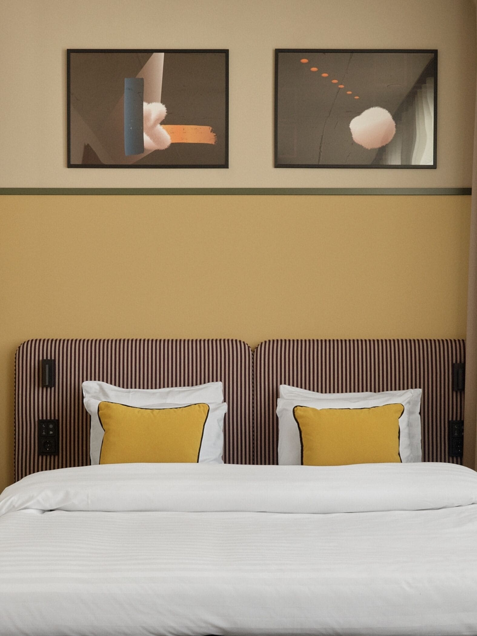A hotel room with a bed, two pillows, a striped headboard, and two abstract paintings on the wall.