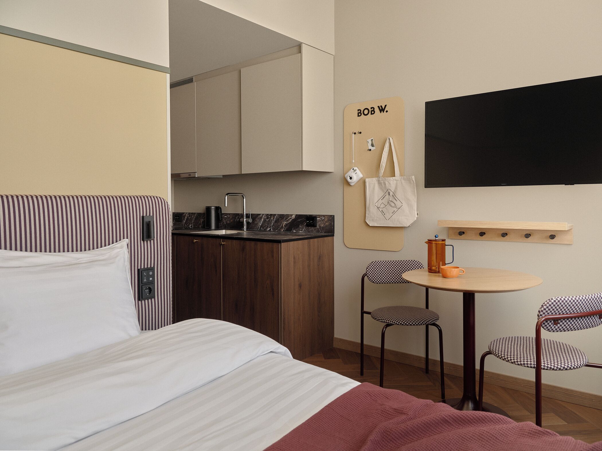 Ein kleines Hotelzimmer mit Bett, einer kleinen Küchenzeile, einem runden Tisch mit Stühlen und einem an der Wand befestigten Fernseher.
