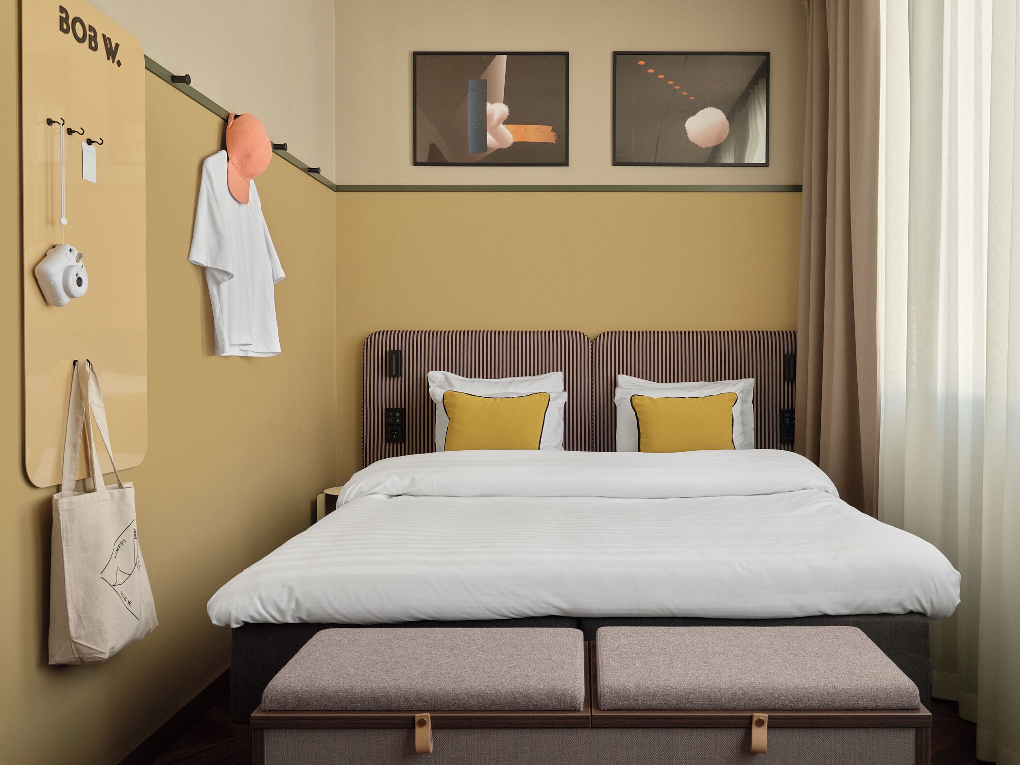 A hotel room with a bed, two pillows, a headboard, a wall-mounted camera, a towel rack, a bag, and artwork.