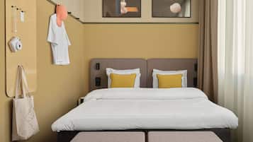 A hotel room with a bed, two pillows, a headboard, a wall-mounted camera, a towel rack, a bag, and artwork.