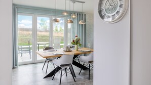 A dining area with a large window, a wooden table, white chairs, hanging pendant lights, and a wall-mounted clock. - Comfortable vacation home Neukamp 17C Seehütte in an idyllic location near Putbus (Putbus)