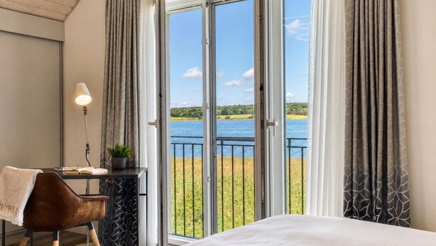 A bedroom with a bed, a desk with a lamp, and a view of a lake through large glass doors. - Comfortable vacation home Neukamp 17C Seehütte in an idyllic location near Putbus (Putbus)