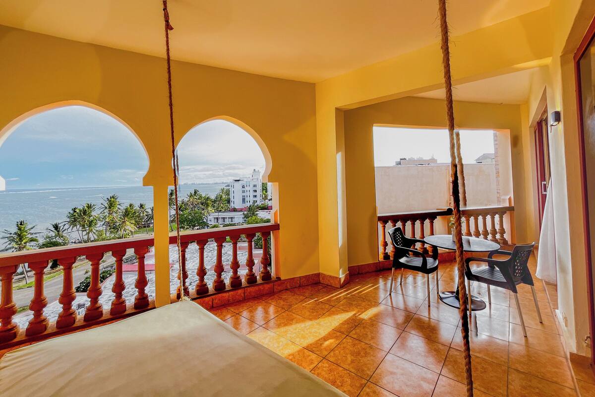 Un balcón con vista al mar y palmeras, una mesa con sillas y una hamaca.