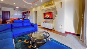 A living room with a blue sofa, a glass coffee table with a fish design, a ceiling fan, a TV with a fireplace display, and a kitchen in the background. - Kuzuri Kwetu Beachfront Apartments (Mombasa)