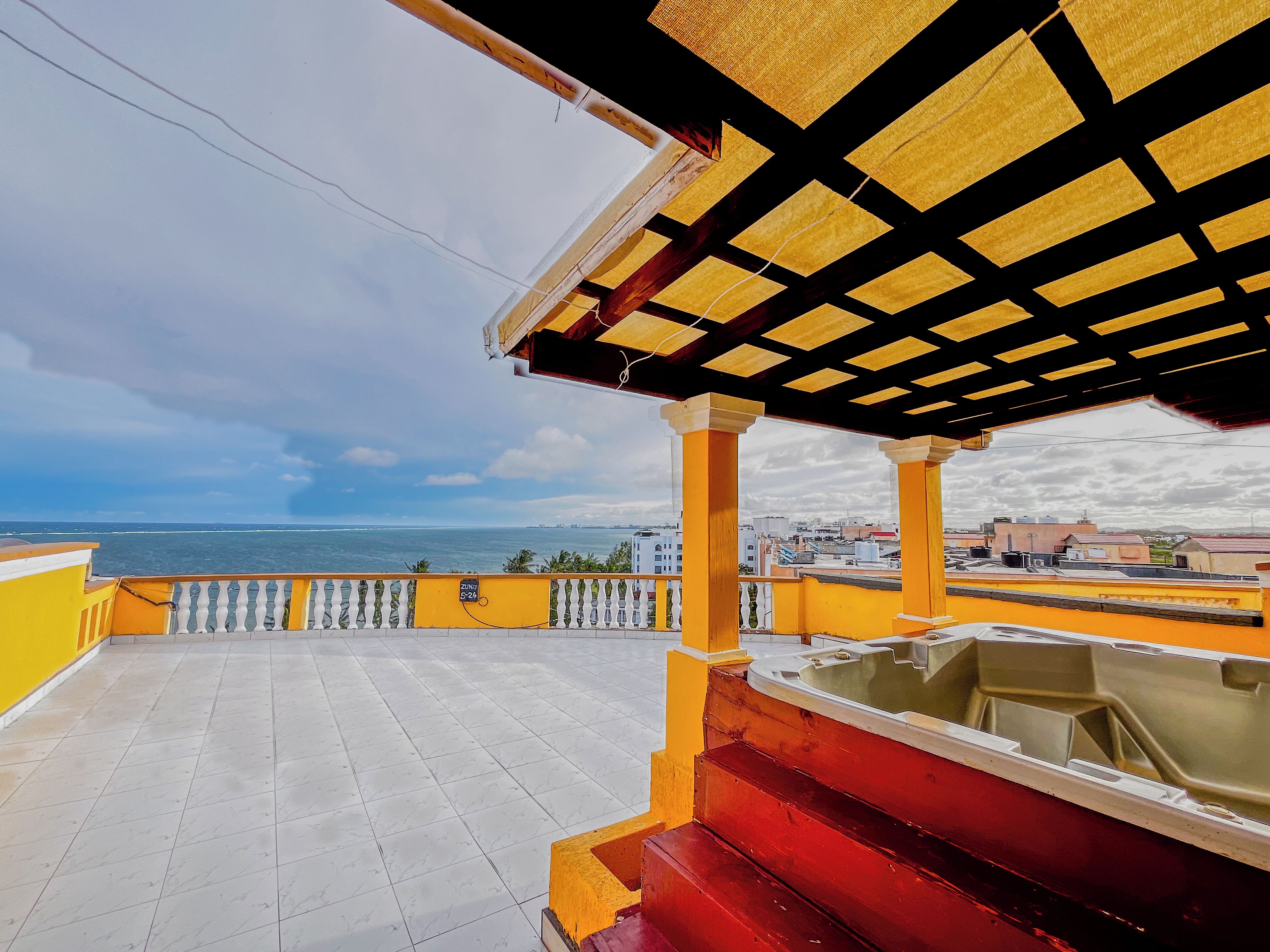 Terraza en la azotea con vista al mar, que presenta una estructura amarilla y roja, suelo de baldosas blancas y una pérgola con diseño negro y amarillo.
