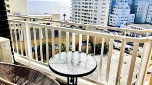 A balcony with wicker chairs and a glass table, overlooking a parking area and buildings with a view of the ocean.