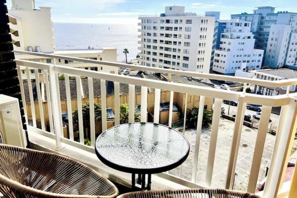 A balcony with wicker chairs and a glass table, overlooking a parking area and buildings with a view of the ocean.