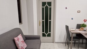 A living room with a grey sofa, a small table with chairs, a green-framed door, and a white wall with a power outlet. - Isabel house (Napoli)