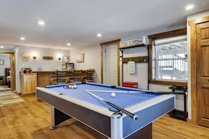 A pool table in a room with wooden flooring, a sign indicating 'SKI SLOPES,' and a 'WELCOME' sign on the wall.