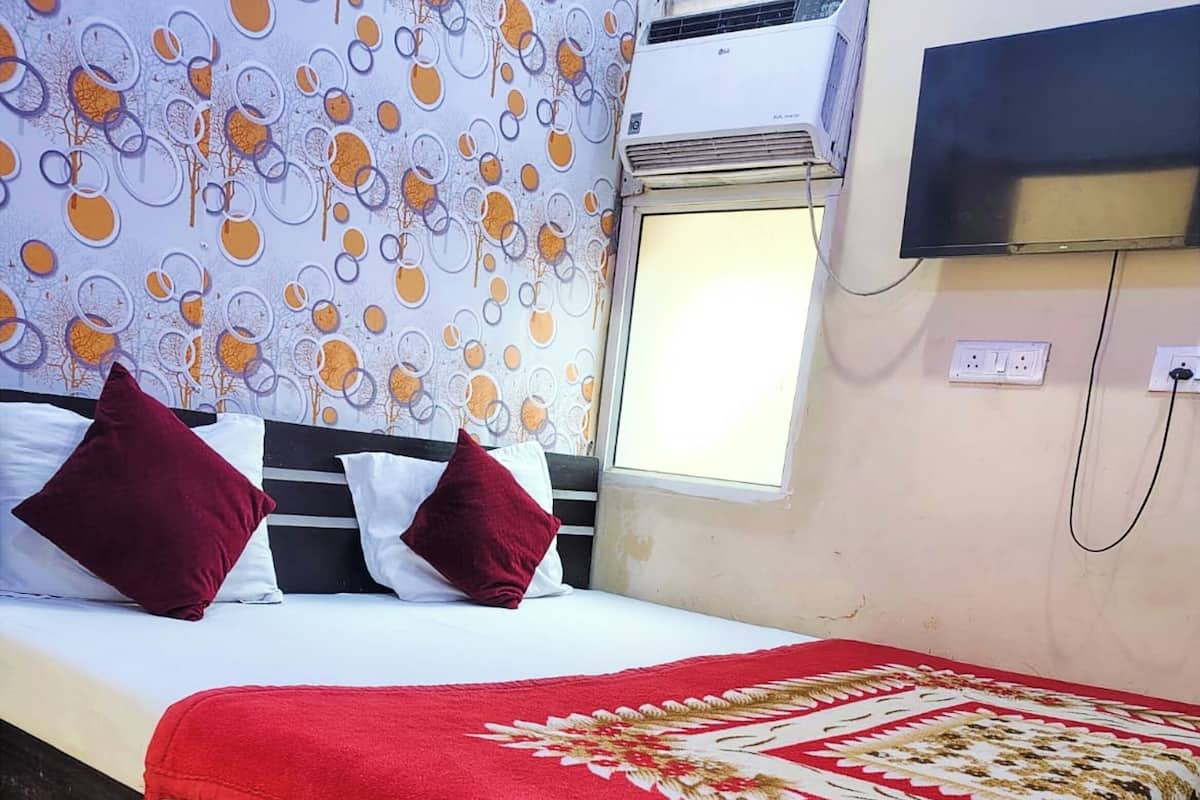 Una habitación de hotel con cama, almohadas, una manta roja, televisor y ventana.
