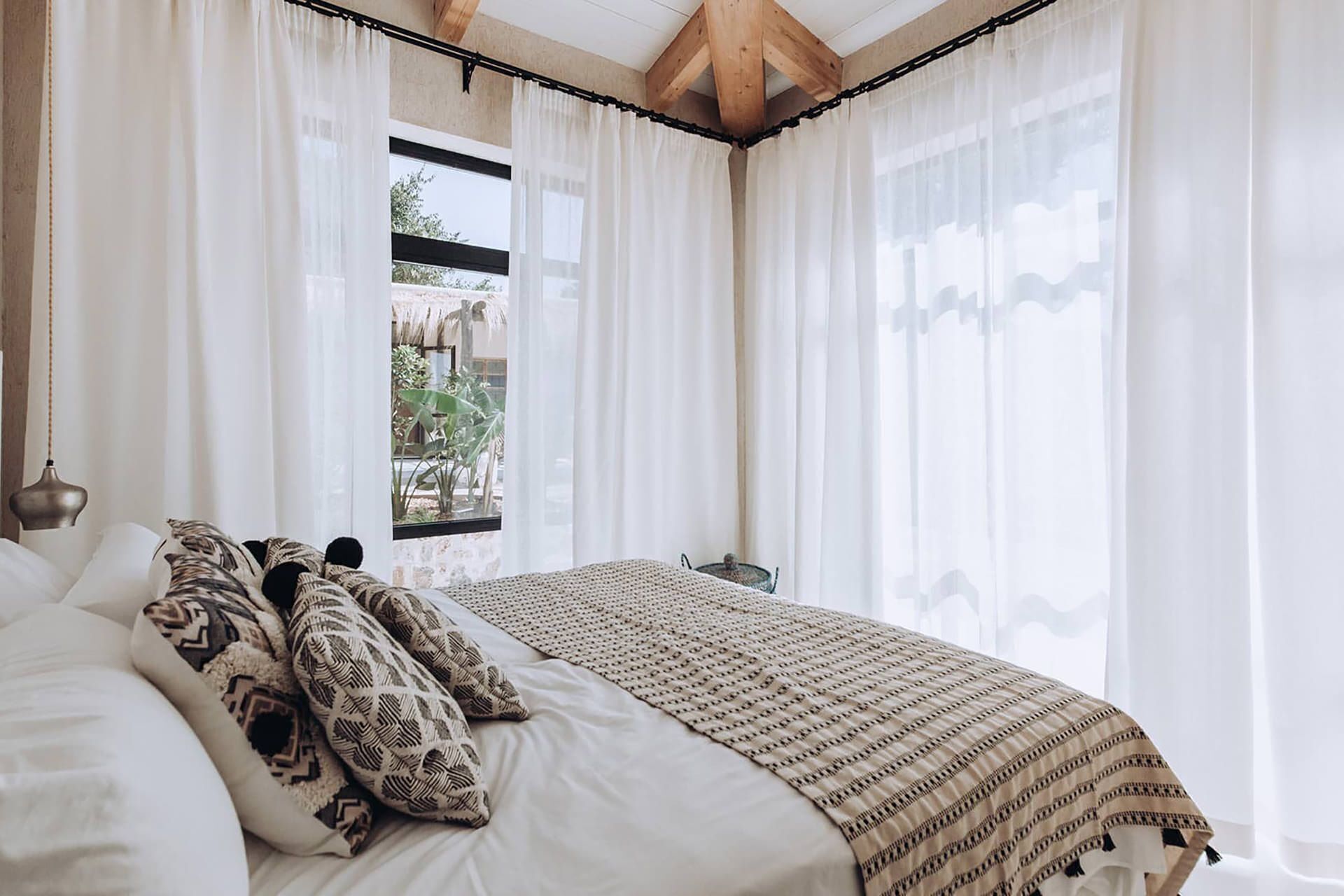 A bedroom with a bed, white curtains, a window, and a view of greenery outside.