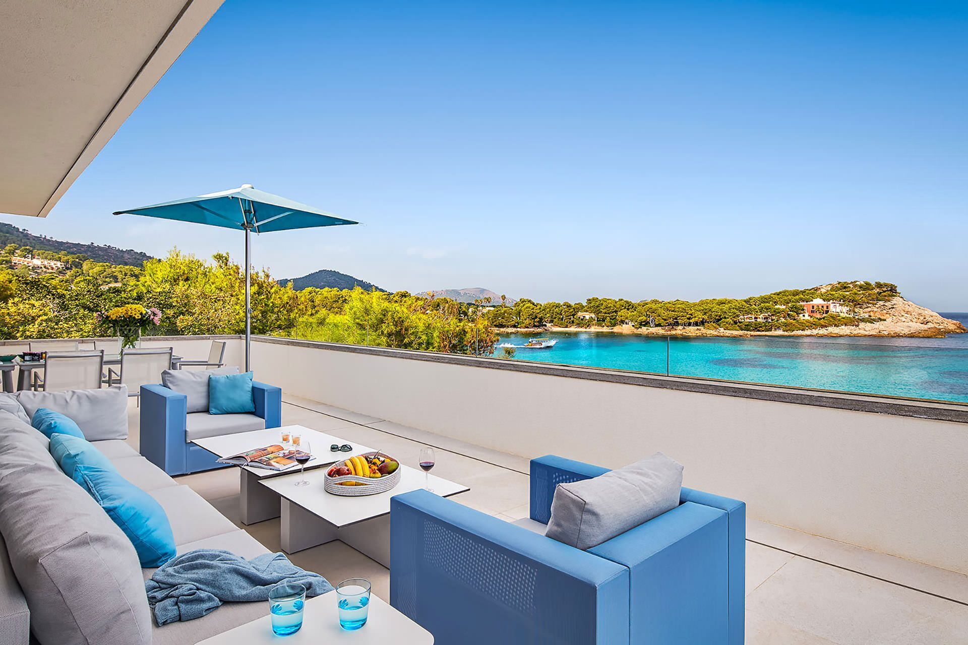 A terrace with a sofa, a table, and an umbrella overlooking a coastal view with a body of water and greenery.