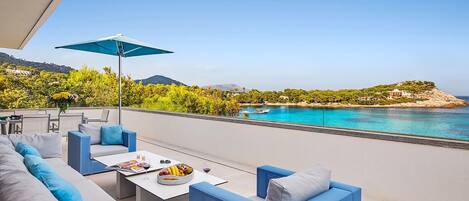 A terrace with a sofa, a table, and an umbrella overlooking a coastal view with a body of water and greenery.