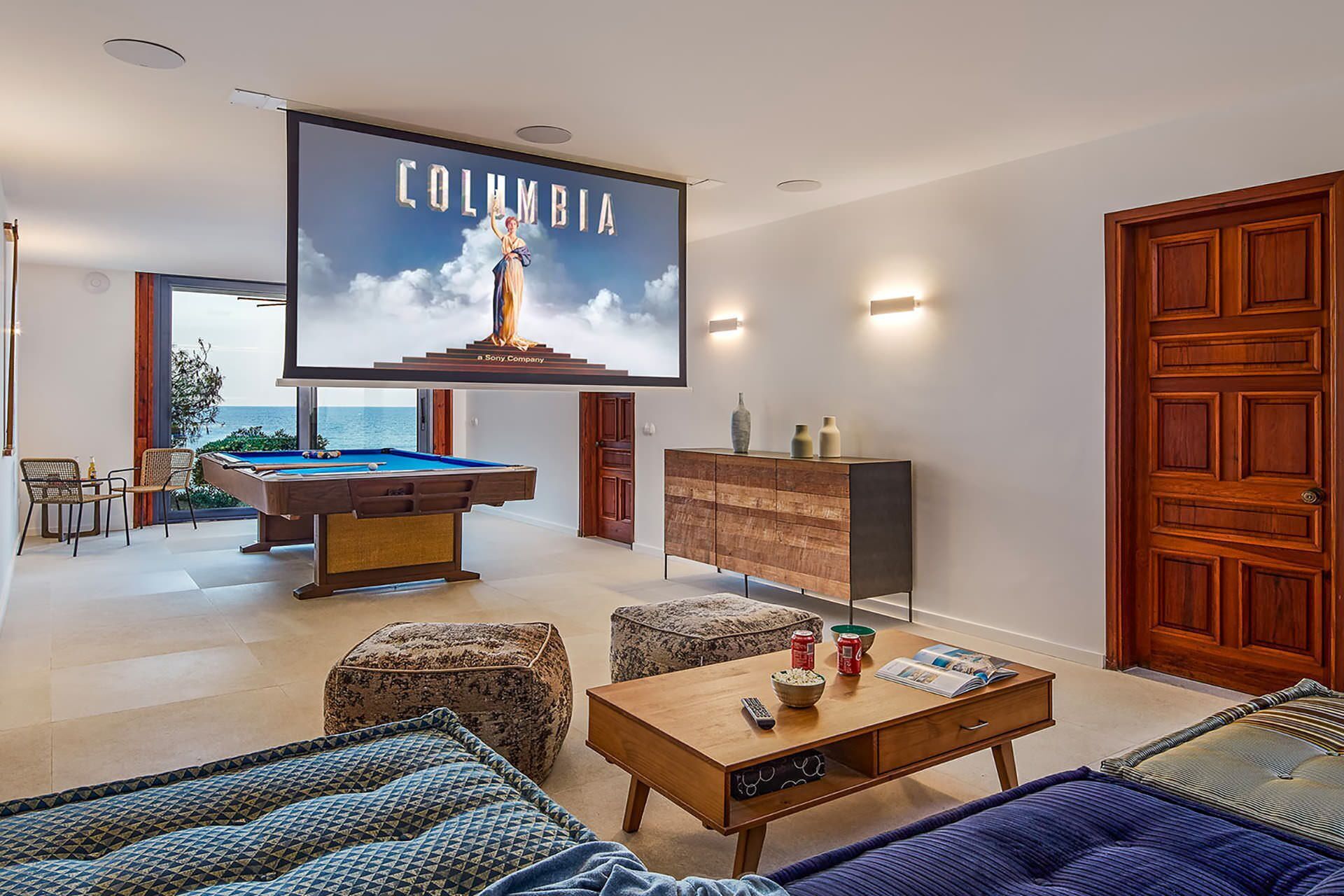A modern living room with a pool table, comfortable seating, a large TV screen displaying 'COLUMBIA', and a view of the sea.