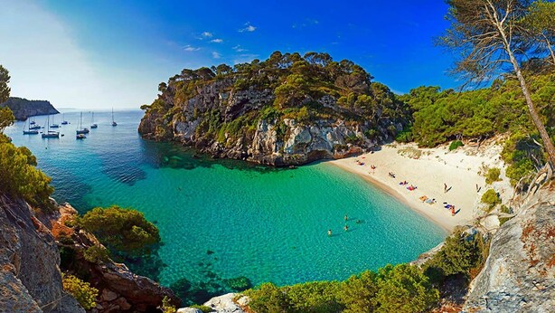 Una playa costera con aguas turquesas cristalinas, un acantilado rocoso y una playa de arena con gente.