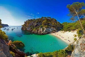 Una playa costera con aguas turquesas cristalinas, un acantilado rocoso y una playa de arena con gente.