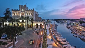 A historic building with a clock tower, a marina with boats, and a sunset sky. - My Rental Homes - Villa Mosaic with private pool, gazebo and barbeque (Mercadal)